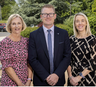 Three adults dressed in business casual attire stand outdoors on a paved area with greenery and parked cars in the background, smiling at the camera. Description generated by AI