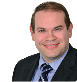 A person with short hair wearing a dark suit jacket, a blue striped shirt, and a striped tie is smiling against a white background. Description generated by AI
