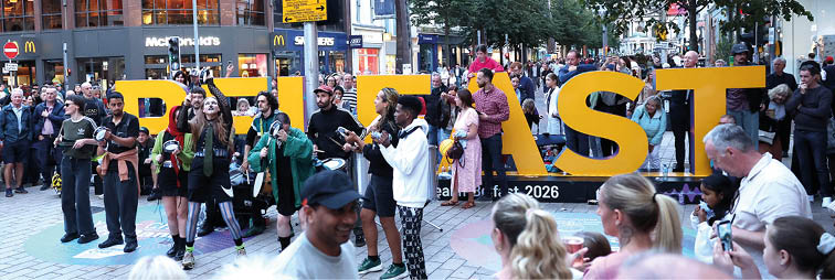 A crowd of people gathers outdoors around large yellow letters spelling “RUTST" with some individuals taking photos and others watching the scene in an urban setting. Description generated by AI