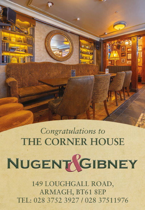 A cozy pub interior with a long cushioned bench, several chairs around wooden tables, warm lighting, and a round mirror on the wall above a sign congratulating The Corner House Nugent & Gibney with an address and phone numbers. Description generated by AI