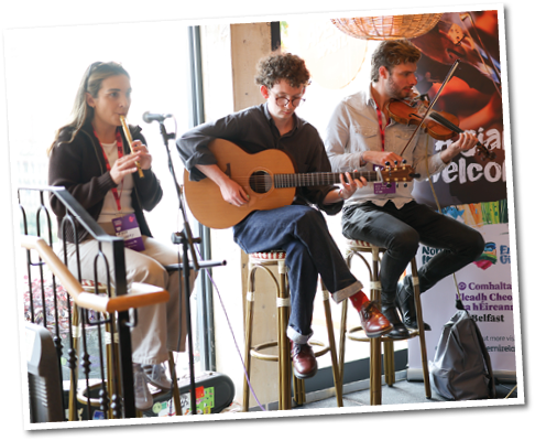 Three adults are playing musical instruments including a flute, guitar, and violin while seated on stools in a cozy indoor setting with hanging wicker lamps and music posters on the walls. Description generated by AI
