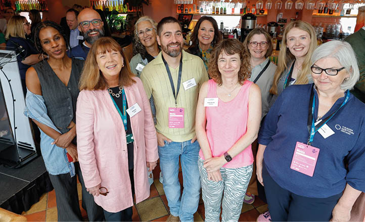 A group of smiling adults wearing conference badges pose together in a warmly lit indoor setting with shelves of bottles and hanging glasses in the background. Description generated by AI
