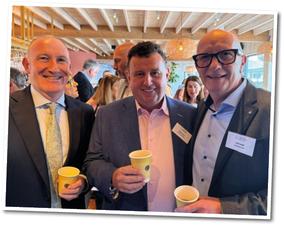 Three adults dressed in business attire are smiling and holding cups while standing closely together in a well-lit indoor setting with other people and hanging lights in the background. Description generated by AI