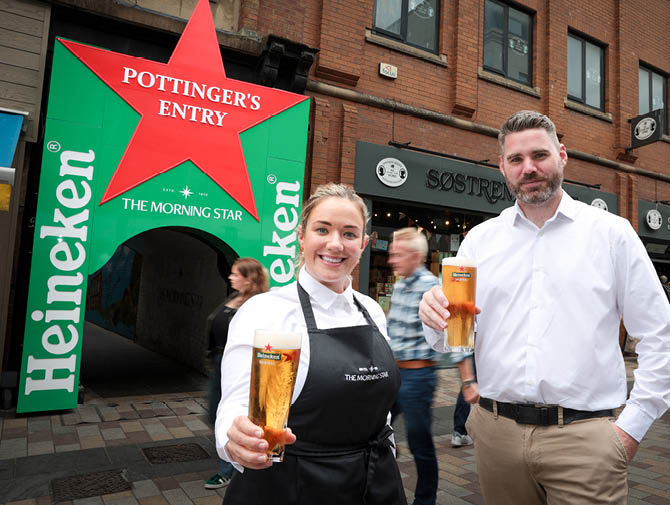 Two adults smiling and holding glasses of beer while standing on a street in front of a large green and red Heineken sign shaped like a star. Description generated by AI