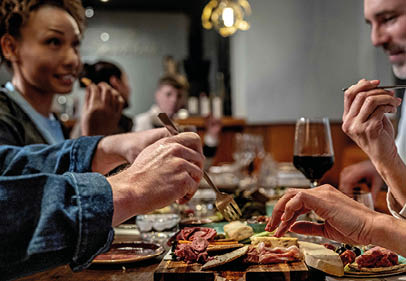 People sitting around a wooden table enjoying a meal with various appetizers and a glass of red wine in a cozy indoor setting. Description generated by AI