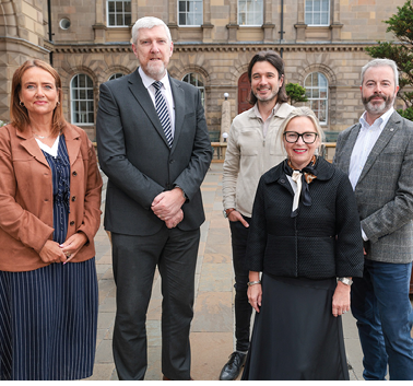 Five adults dressed in business and casual attire stand together outdoors with a stone pavement background. Description generated by AI
