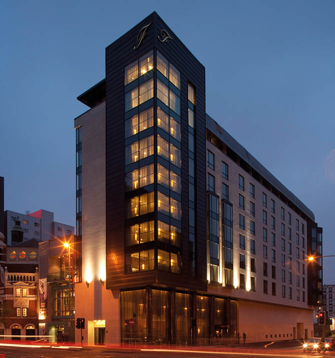 A modern multi-story building with illuminated windows stands at a busy urban intersection during dusk with light trails from passing vehicles. Description generated by AI
