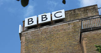 A brick building with a large BBC sign mounted near the top against a blue sky with some tree leaves visible at the edges. Description generated by AI