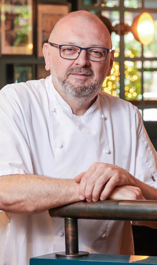 A person wearing glasses and a white chef's coat is leaning on a railing in a warmly lit indoor setting. Description generated by AI