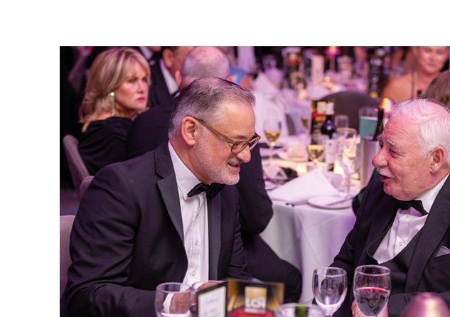 Two adults dressed in formal attire are engaged in conversation while seated at a table set with glasses, menus, and a small lamp in a banquet or event setting. Description generated by AI