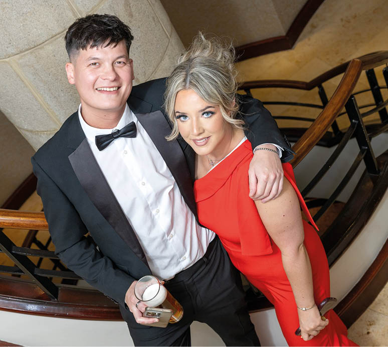 Two adults dressed in formal attire stand closely together on a staircase, with one holding a drink and both smiling. Description generated by AI