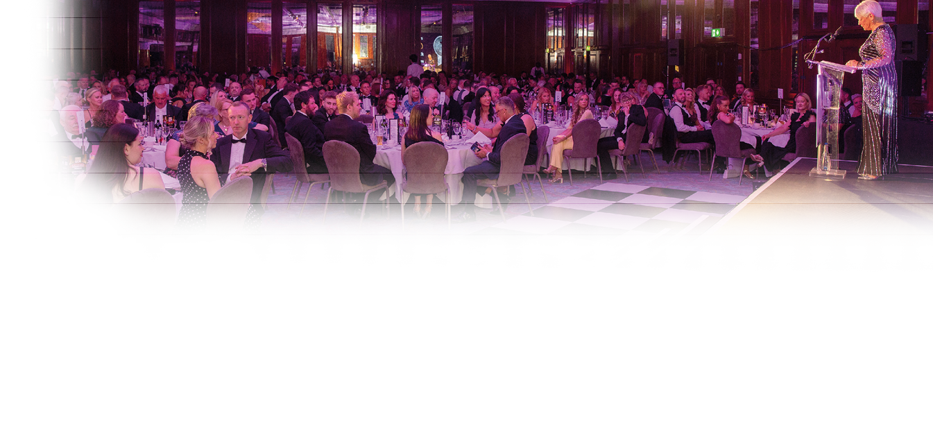A large group of people dressed in formal attire are seated at round tables in a grand hall while a person in a sparkly dress speaks at a podium on a stage. Description generated by AI