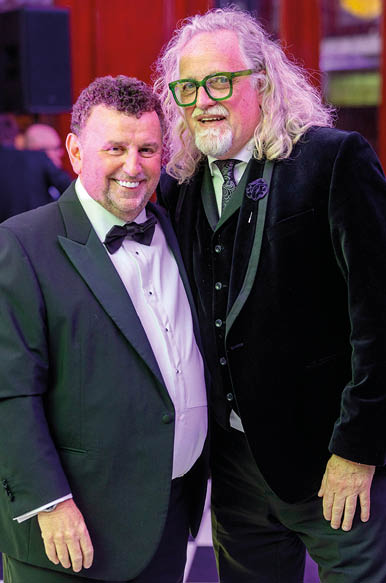 Two adults dressed in formal attire are standing close together and smiling in an indoor setting with warm lighting. Description generated by AI