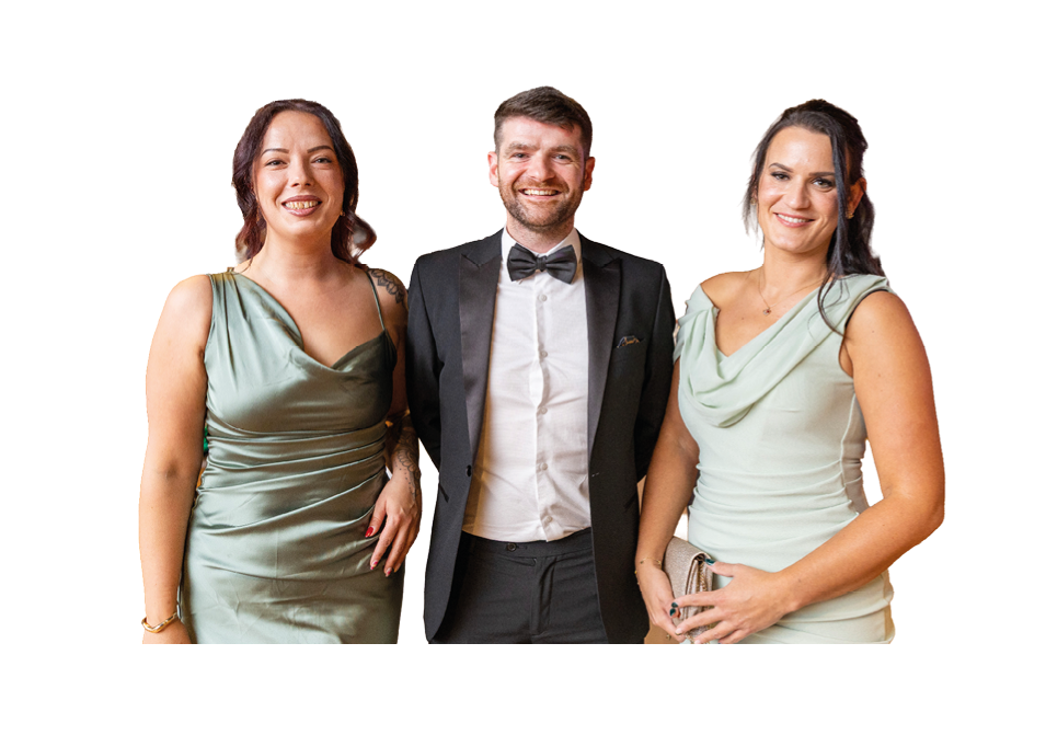 Three adults dressed in formal attire stand closely together smiling against a white background. Description generated by AI
