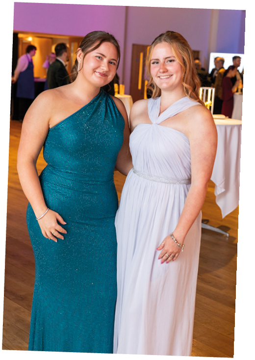 Two adults dressed in formal gowns are standing close together and smiling in an indoor event setting. Description generated by AI