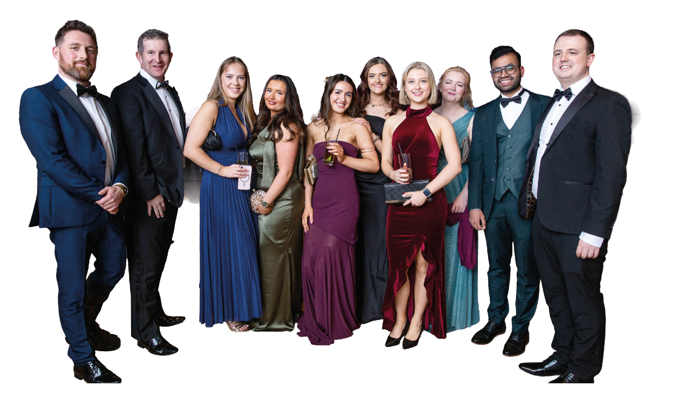 A group of adults dressed in formal evening attire stand closely together smiling against a white background. Description generated by AI