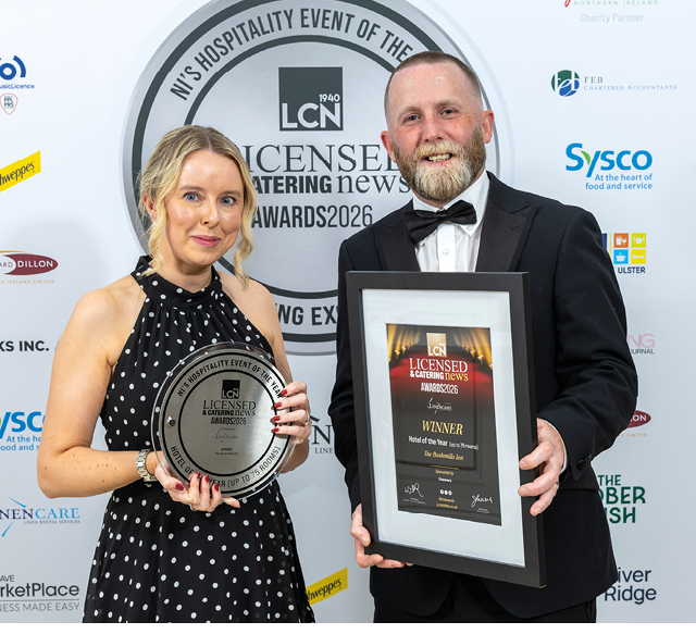 Two adults dressed formally hold an award plaque and a framed certificate while standing in front of a backdrop featuring logos and text related to a hospitality event. Description generated by AI