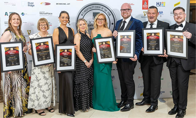 Seven adults dressed in formal attire stand side by side holding framed awards in front of a backdrop with various logos at a hospitality event. Description generated by AI