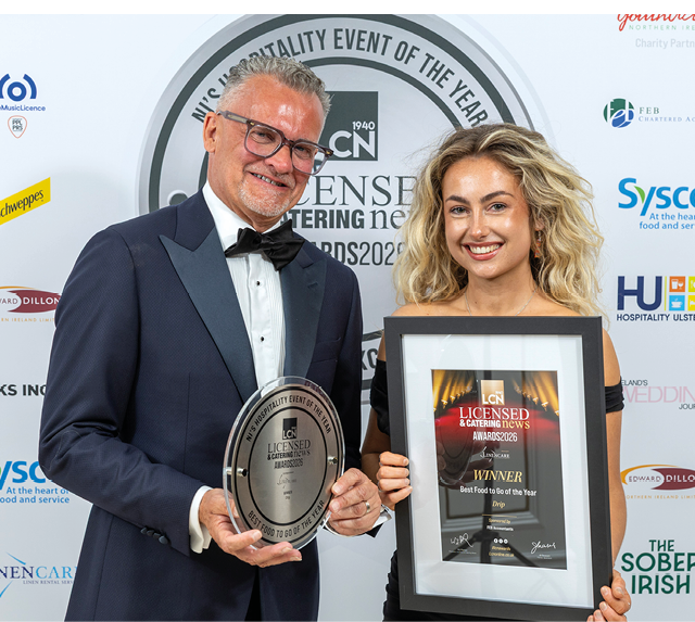 Two adults dressed formally are smiling and holding framed awards at a licensed catering news event. Description generated by AI