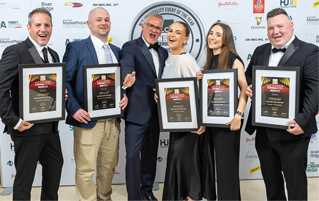 Five adults dressed in formal attire stand smiling and holding framed awards against a backdrop with various logos. Description generated by AI