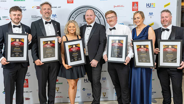 Seven adults dressed in formal attire stand in a row holding framed awards against a backdrop with various logos. Description generated by AI