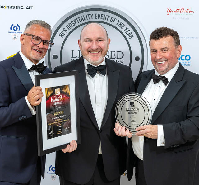 Three adults in tuxedos smile while holding framed awards at a formal hospitality event. Description generated by AI