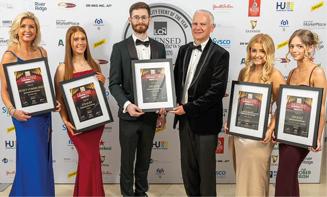 Five adults dressed in formal evening attire stand in a row holding framed awards against a backdrop with various sponsor logos. Description generated by AI