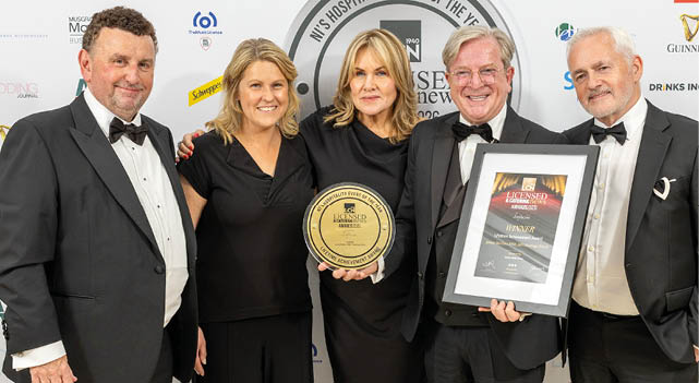 Five adults dressed in formal attire stand together smiling, with one holding a framed award and another holding a round plaque, in front of a backdrop featuring various logos. Description generated by AI