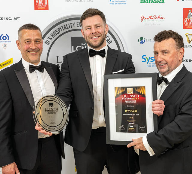 Three adults in tuxedos smiling and holding an award plaque and a framed certificate at a formal event with a branded backdrop. Description generated by AI