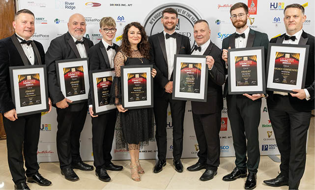 Seven adults dressed in formal attire stand side by side holding framed awards and smiling in front of a backdrop with various logos. Description generated by AI