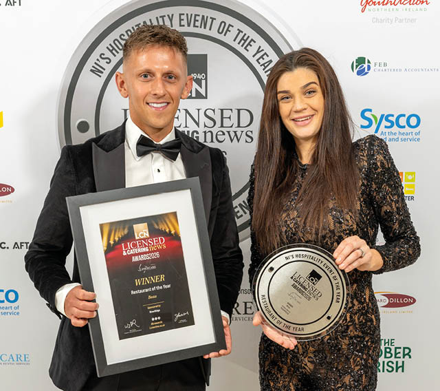 Two adults dressed in formal attire smile while holding framed awards at a hospitality event. Description generated by AI