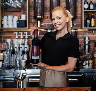 Wine tasting and serving in the winery. Front view of a female wine connoisseur in a modern uniform working behind the bar. Woman holds a bottle of fine wine with both hands and smiles. Wine promotion