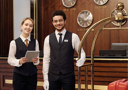 Happy young elegant receptionist and bellboy standing in lounge of luxurious hotel and looking at camera while waiting for new guests