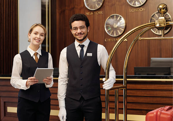 Happy young elegant receptionist and bellboy standing in lounge of luxurious hotel and looking at camera while waiting for new guests