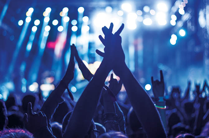 Audience with hands raised at a music festival and lights streaming down from above the stage. Soft focus, blurred movement.