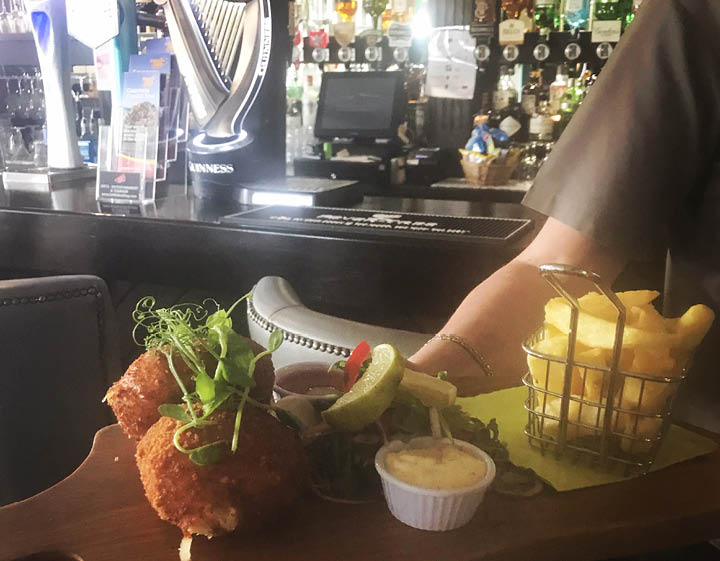 A wooden tray with a sandwich, pickles, and potato chips. Description generated by AI