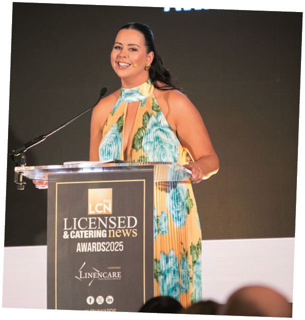 A woman in a flowered dress is speaking at an awards ceremony. Description generated by AI