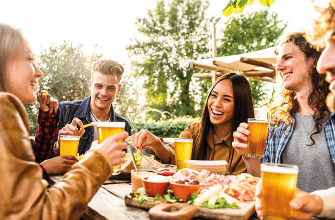 Group of multi ethnic friends having backyard dinner party together - Diverse young people sitting at bar table toasting beer glasses in brewery pub garden - Happy hour, lunch break and youth concept