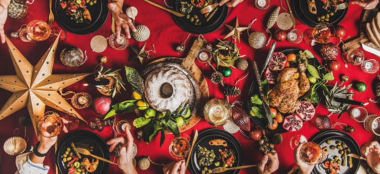 Friends celebrating Christmas. Flat-lay of people eating and talking over festive table with red cloth with champagne, roasted chicken, bundt cake, fruits, decorations, top view, wide composition