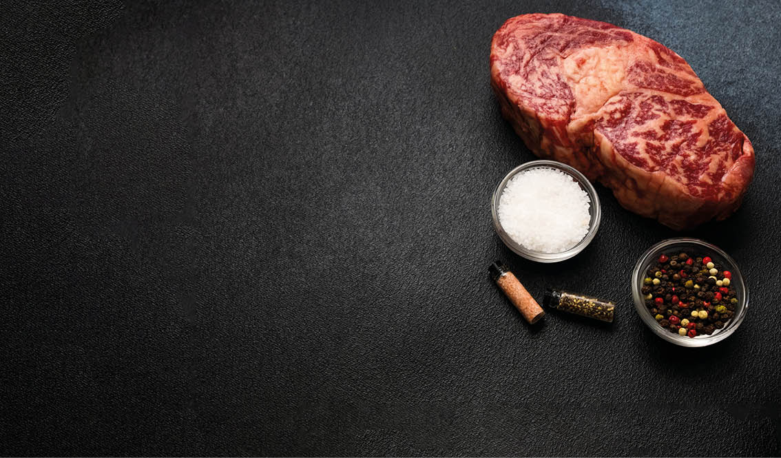 A marbled beef steak rests on a dark surface accompanied by bowls of colorful peppercorns and salt. Two small spice containers also sit nearby ready for seasoning before cooking.