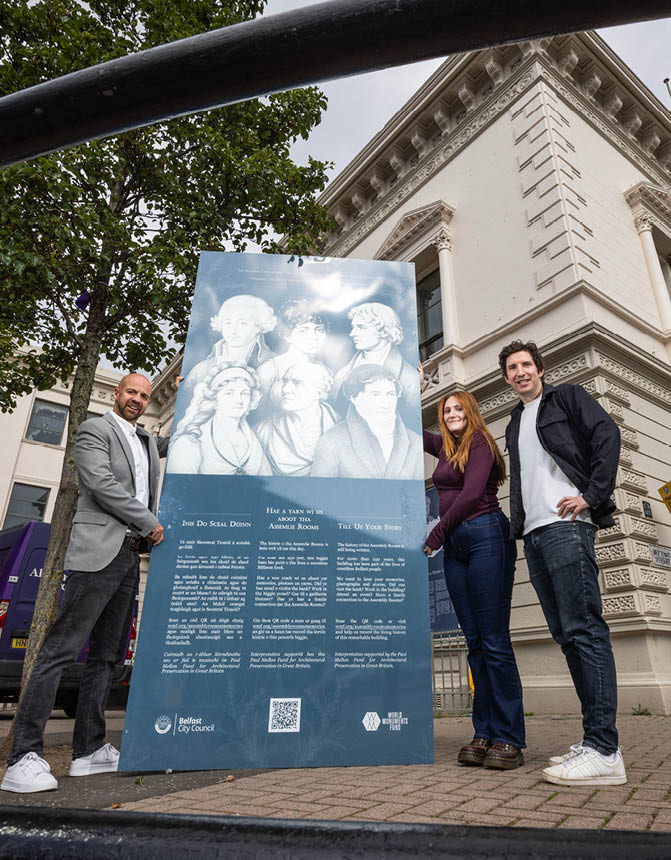 Three adults stand outdoors on a sidewalk holding a large informational sign with historical portraits and text in front of a classical-style building. Description generated by AI