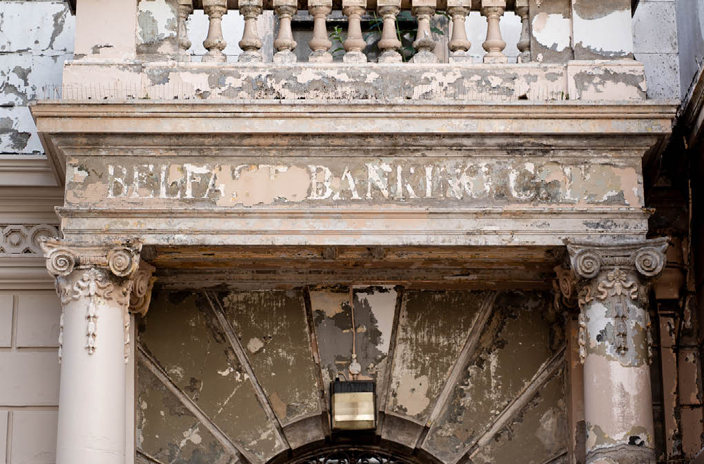 A weathered stone entrance with ornate columns and a faded sign reading “BELFAST BANKING CO." above a peeling archway. Description generated by AI