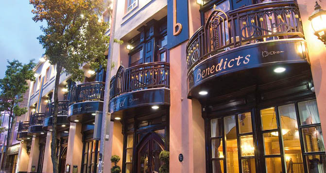 A warmly lit street scene at dusk shows a row of buildings with balconies and signs, including one prominently displaying the name Benedicts. Description generated by AI