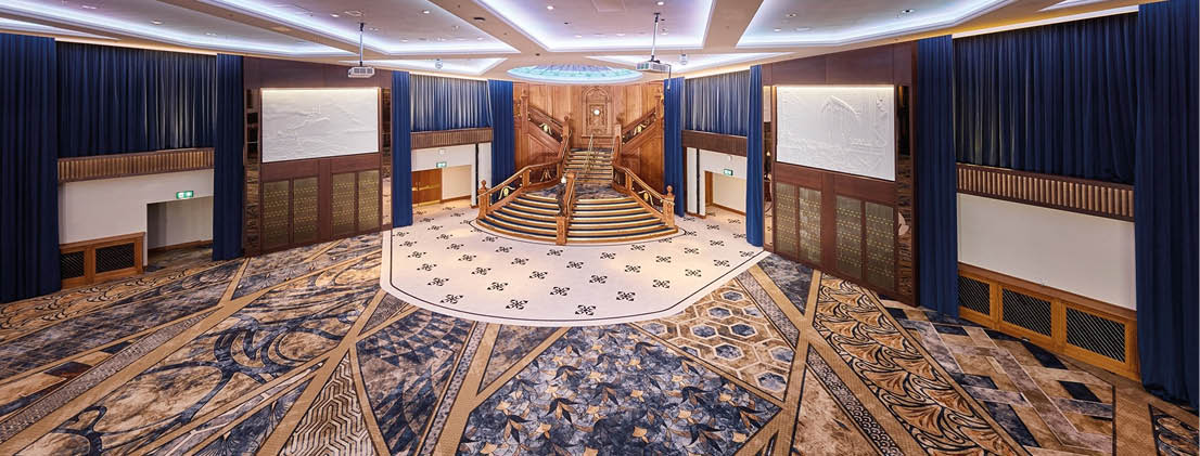 A large, ornate room with patterned carpet, dark blue curtains, wooden paneling, and a central raised platform with steps and a wooden backdrop. Description generated by AI