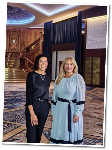Two adults smiling and standing side by side in a spacious, elegant interior with a grand staircase and decorative carpet, one wearing a black outfit and the other wearing a light blue dress with dark accents. Description generated by AI