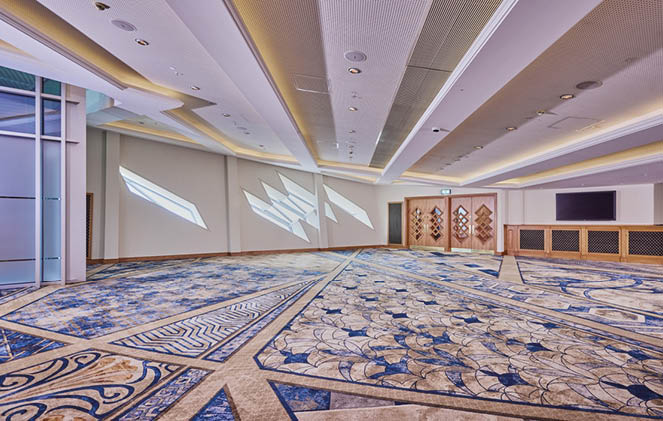 A spacious, modern conference room with patterned blue and beige carpet, recessed ceiling lights, geometric wall windows, wooden double doors, and a large flat-screen TV mounted on the wall. Description generated by AI