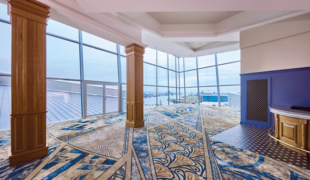 A spacious room with large floor-to-ceiling windows, wooden columns, and a patterned carpet overlooking a waterfront view. Description generated by AI