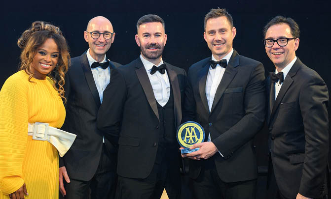 Five adults dressed in formal attire stand together smiling, with one holding a circular award featuring a blue and yellow design. Description generated by AI