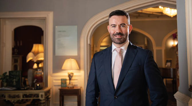 A person wearing a dark suit and light-colored tie is standing and smiling in a warmly lit, elegant room with classic furniture and lamps. Description generated by AI