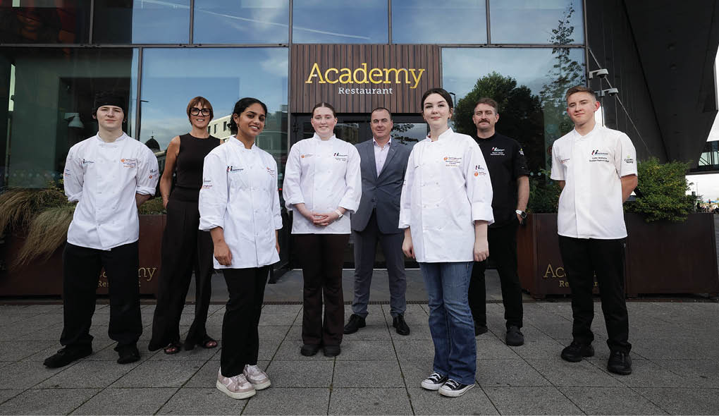 Seven people, including five wearing chef jackets and two in business attire, stand in front of a building with a sign that reads “Academy Restaurant." Description generated by AI